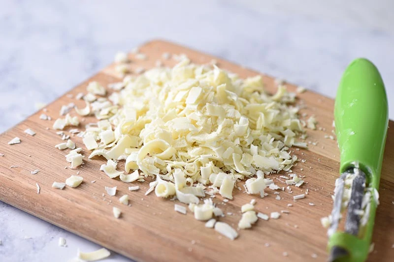 shredded white chocolate for white chocolate raspberry cheesecake with vegetable peeler on wooden cutting board