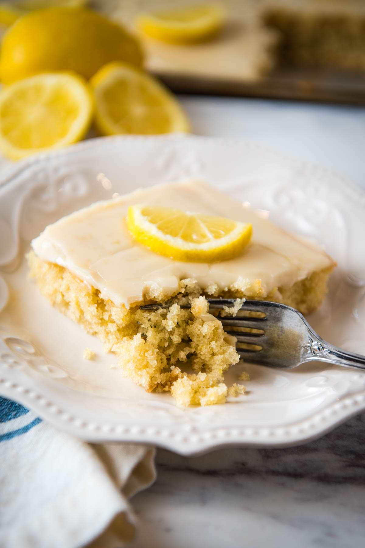 sliced square of lemon Texas sheet cake topped with cooked lemon frosting and a fresh lemon slice and with a bite taken out on a small white saucer
