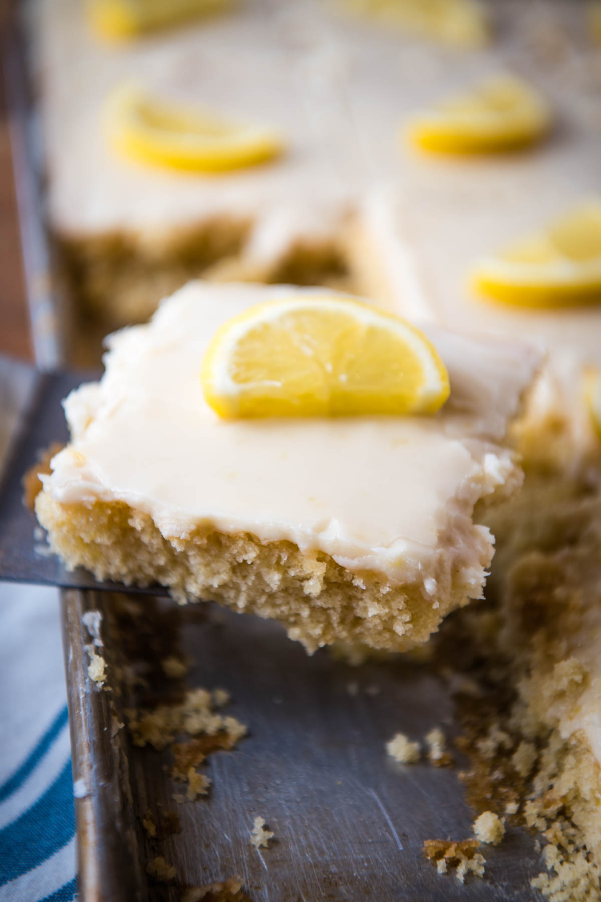 closeup photo of a small silver spatula holding up a sliced square of lemon Texas sheet cake topped with a cooked lemon frosting and a fresh lemon slice over large lemon sheet cake in a jelly roll pan
