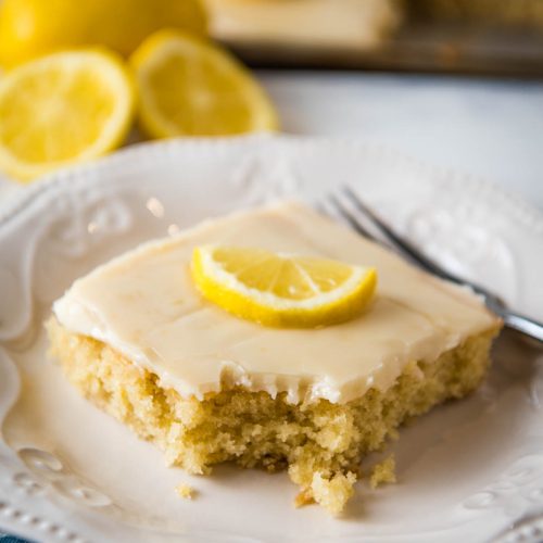 sliced square of lemon Texas sheet cake topped with a cooked lemon frosting and a fresh lemon slice with a bite taken out on a small white saucer