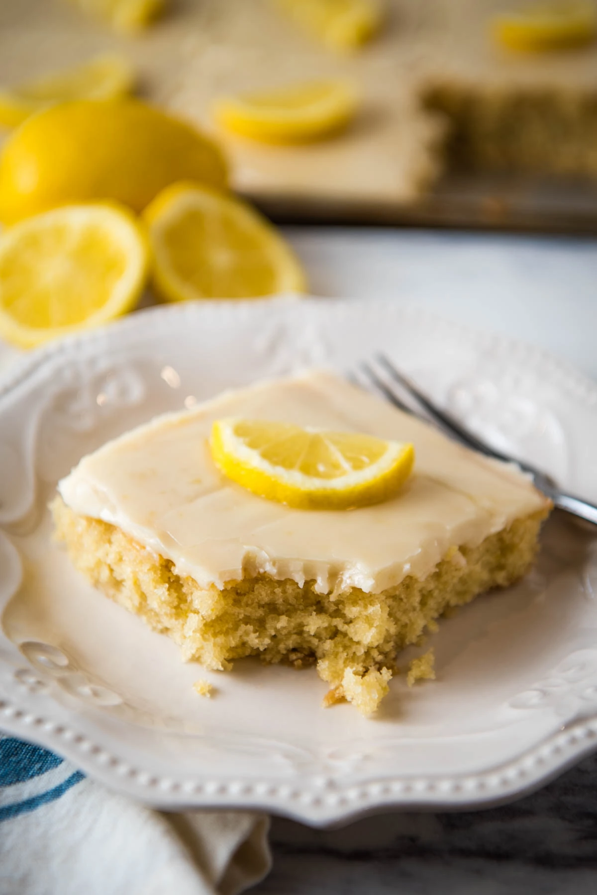 sliced square of lemon Texas sheet cake topped with a cooked lemon frosting and a fresh lemon slice with a bite taken out on a small white saucer 