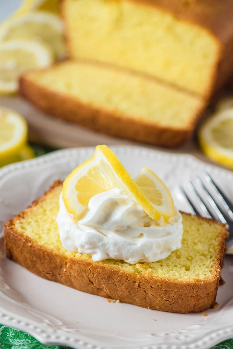 slice of lemon pound cake on white plate with whipped cream and slice of fresh lemon on top of the cake