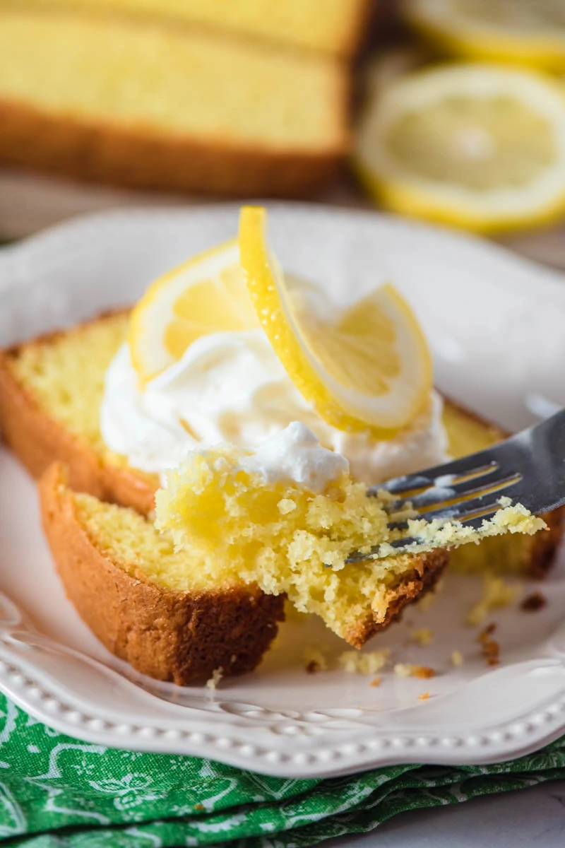 bite of moist lemon pound cake on fork with slice of cake on white plate in background