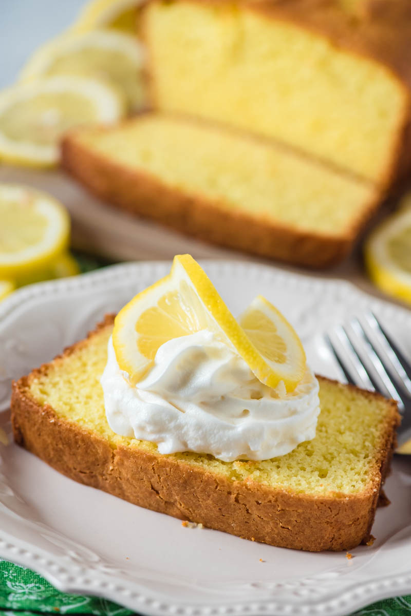 slice of lemon loaf cake on white plate with whipped cream and fresh lemon slice on top