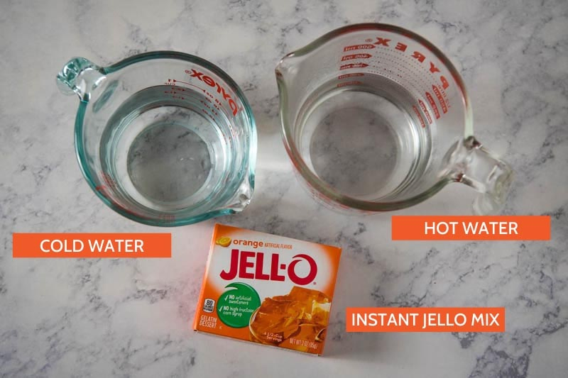 box Jello ingredients, including orange Jello gelatin mix and hot water and cold water in glass measuring cups on white marble countertop