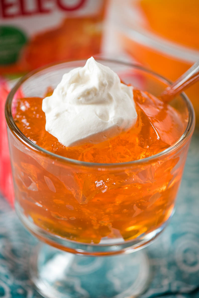glass mini trifle dish full of orange gelatin and topped with Dream Whip whipped cream, with spoon, on blue linen cloth