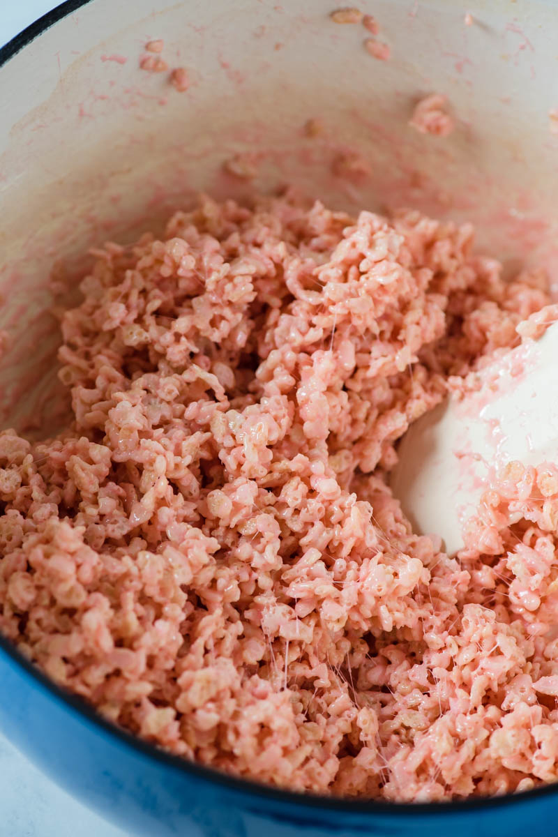 stirring pink colored Rice Krispie treats with white spatula in blue Dutch oven