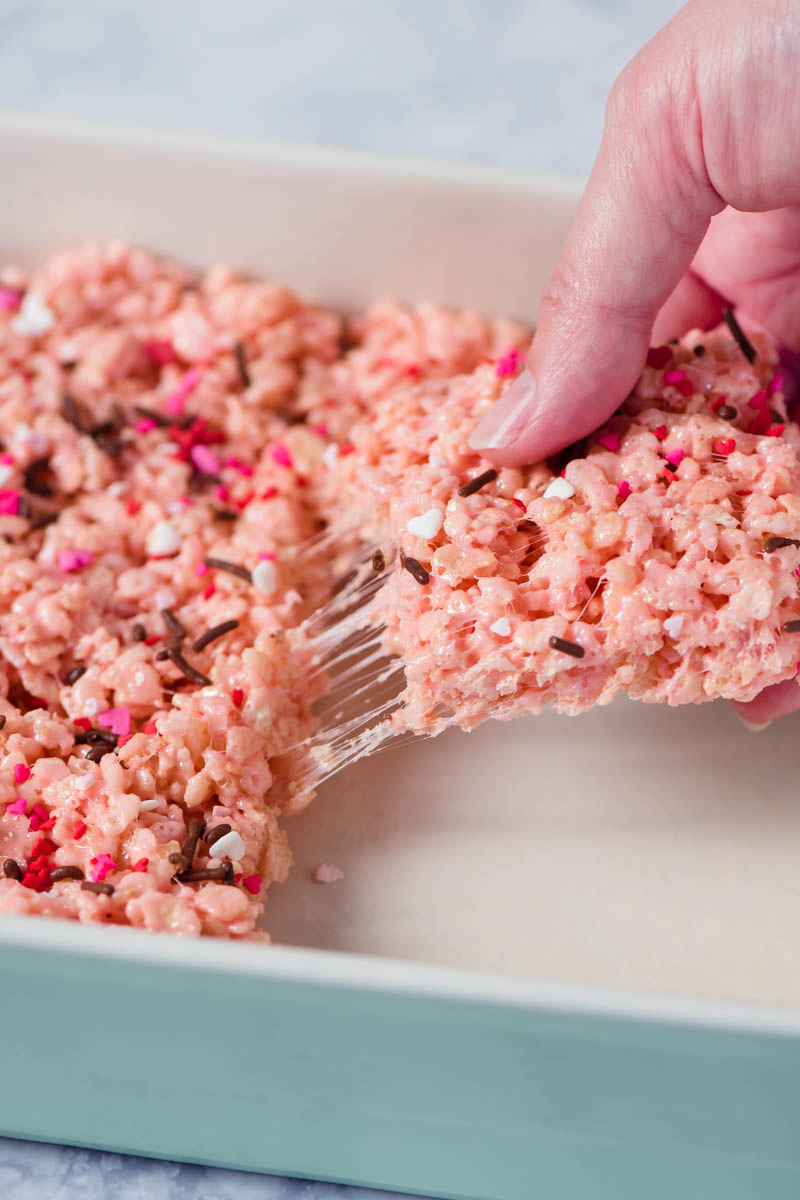 hand pulling pink marshmallow cereal treats from other treats in mint green baking dish, with gooey strings of marshmallow