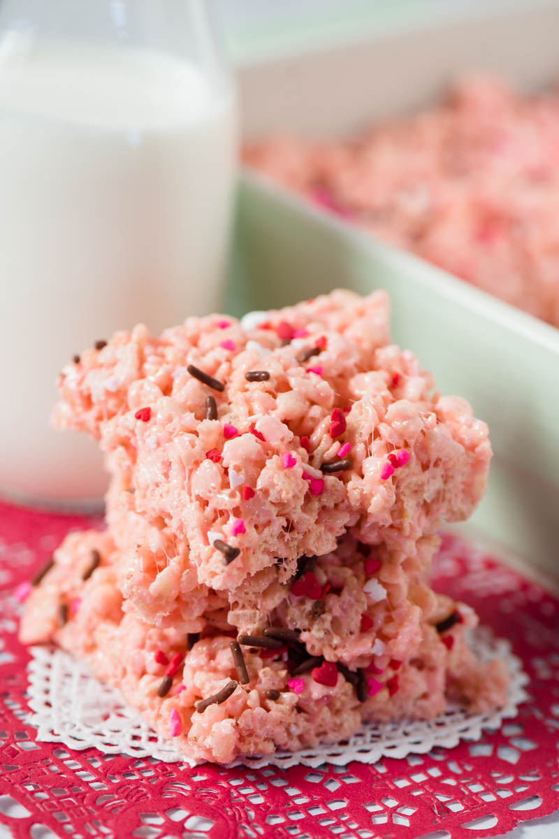 stack of pink Valentine Rice Krispie treats with sprinkles on top, sitting on white and red Valentine paper heart doilies, with mint green baking dish and jar of milk behind