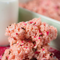 stack of pink Rice Krispie treats with sprinkles, with bite taken, sitting on white and red paper heart, with mint green baking dish and glass of milk behind