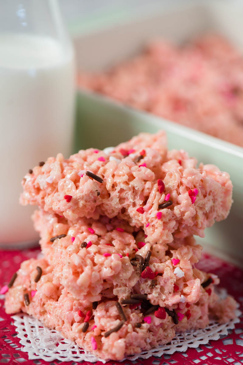stack of sprinkle-topped pink Valentine Rice Krispie treats with bite taken, sitting on white and red Valentine paper heart, with mint green baking dish and glass of milk behind