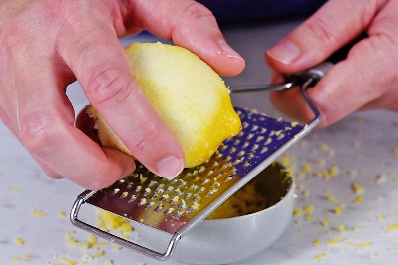 hands zesting lemon with lemon zester tool over small gray bowl