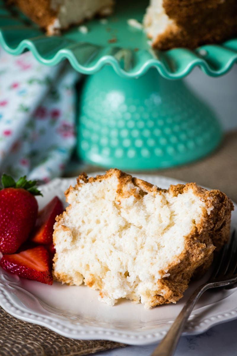 slice of angel cake with strawberries on white plate with fork, with green cake stand behind