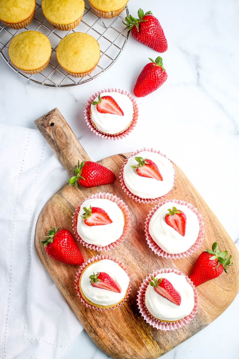 cupcakes with strawberry filling, topped with whipped cream frosting and fresh strawberries, on wooden cutting board, on white marble countertop