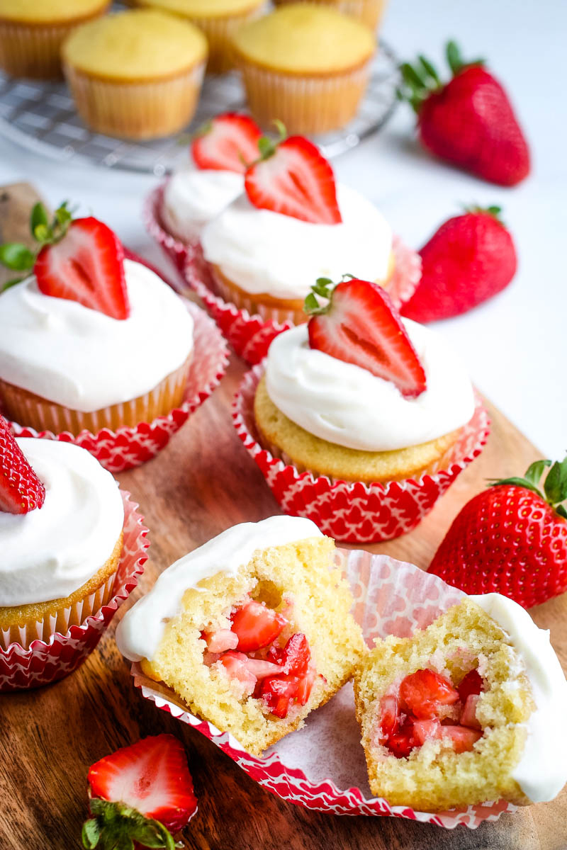vanilla cupcakes filled with fresh strawberries and topped with whipped cream frosting and fresh strawberry slices, in red cupcake liners, on wooden cutting board