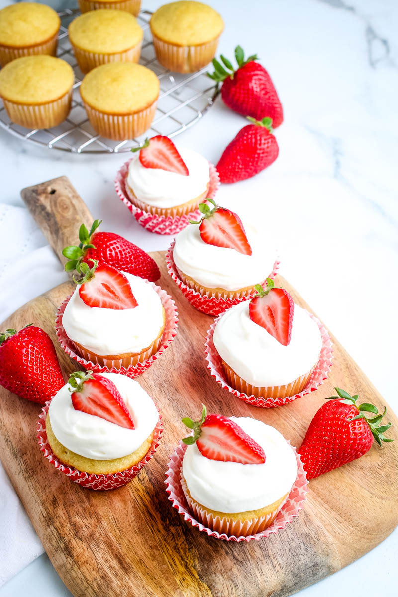 strawberry and whipped cream topped cupcakes sitting in red cupcake liners on wooden cutting board on white marble countertop
