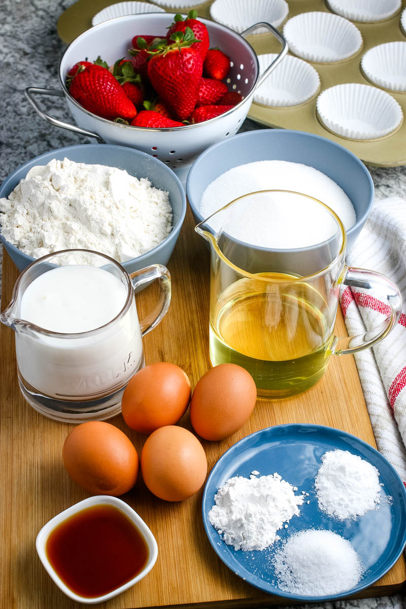 ingredients for shortcake cupcakes, including eggs, vanilla extract, flour, sugar, canola oil, and strawberries in bowls and measuring cups on wooden cutting board