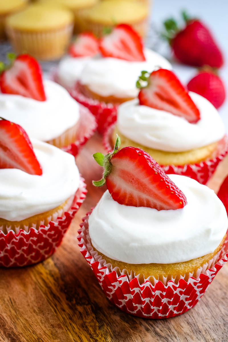 strawberry topped strawberry shortcake cupcakes with whipped cream frosting, in red cupcake liners, on wooden cutting board