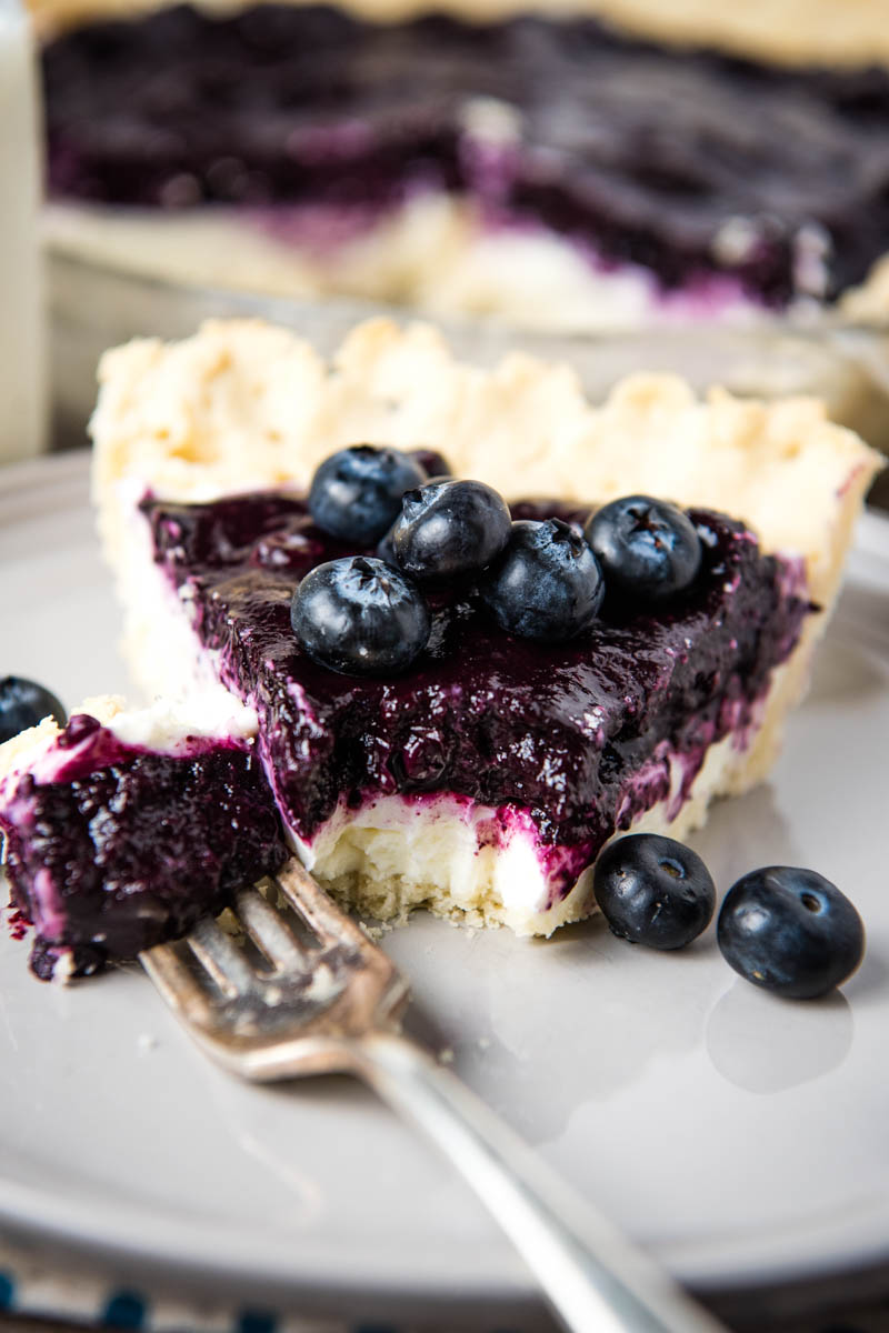 slice of blueberry cream cheese pie with blueberries on top and bite taken out of pie on gray plate with fork and bite