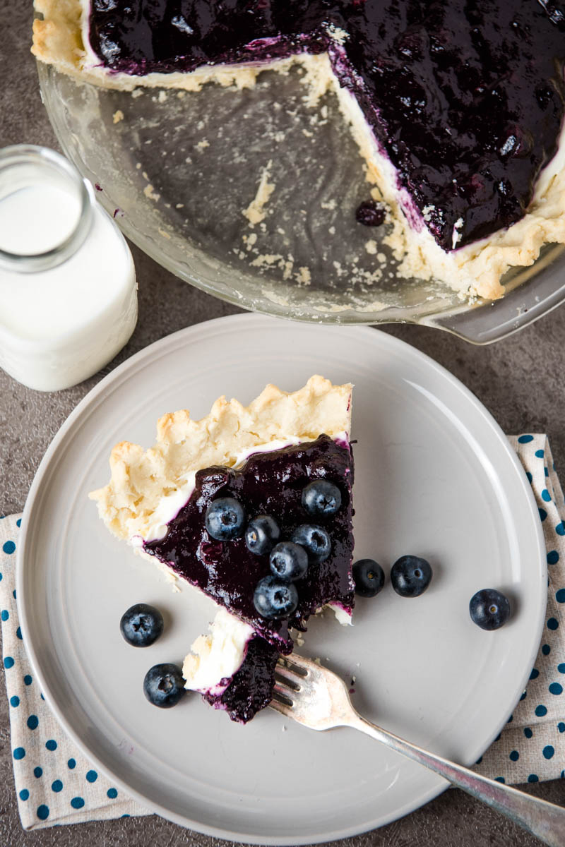 blueberry cream pie sliced and served on gray plate with fresh blueberries and a fork, along with a jar of milk