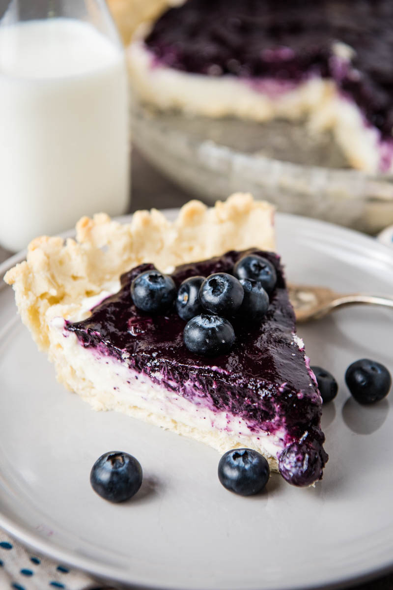 whole slice of blueberry cream cheese pie on gray plate with fork, fresh blueberries, and jar of milk