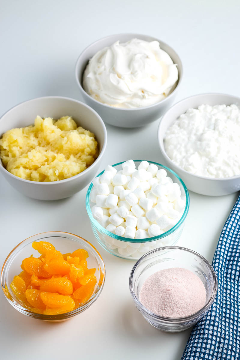 orange Jello salad ingredients, including mandarin oranges, orange Jello mix, mini marshmallows, crushed pineapple, cottage cheese, and whipped cream in small gray bowls on white countertop with blue and white checked cloth
