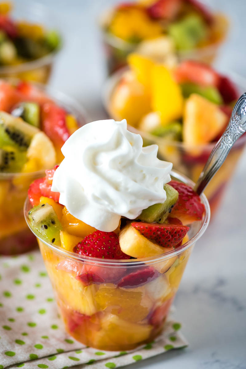 closeup photo of tropical fruit salad cup with dollop of whipped cream on top and a small silver spoon inserted into it