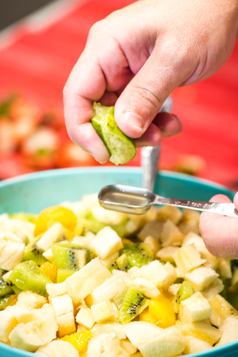 closeup photo of hand-squeezing fresh lime into a silver steel tablespoon over top of homemade fruit salad mixture in a large blue mixing bowl