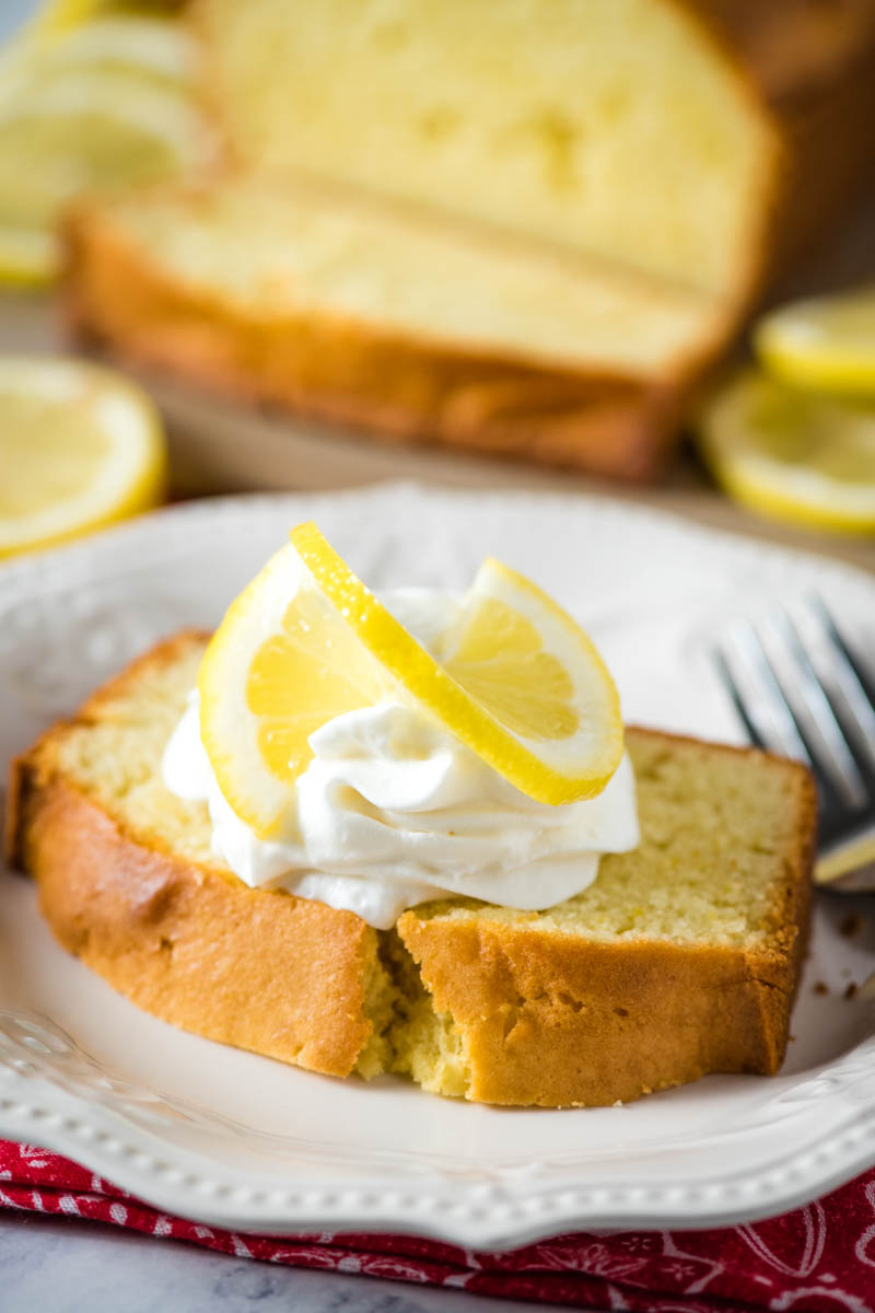 slice of gluten-free lemon loaf, topped with whipped cream and a slice of lemon, on white plate with fork