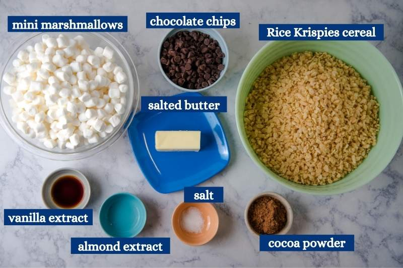 ingredients for chocolate rice crispy treats on white marble countertop, including mini marshmallows, chocolate chips, Rice Krispies cereal, salted butter, vanilla extract, almond extract, salt, and cocoa powder