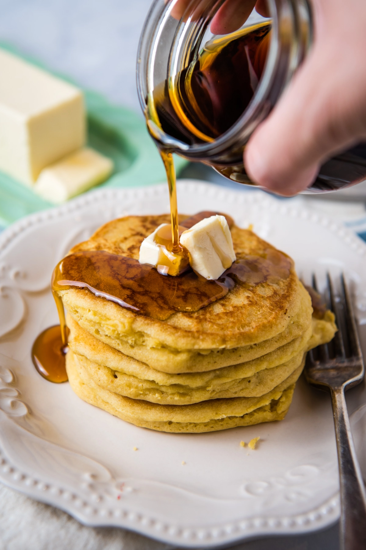 hand pouring maple syrup on stack of gluten-free fluffy pancakes with butter, on white plate with fork