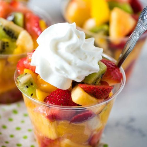 homemade tropical fruit salad cups topped with a dollop of homemade whipped cream on a white and green polka-dot towel