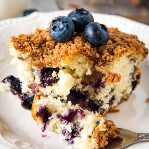 slice of blueberry coffee cake topped with blueberries on white plate and bite of cake on fork