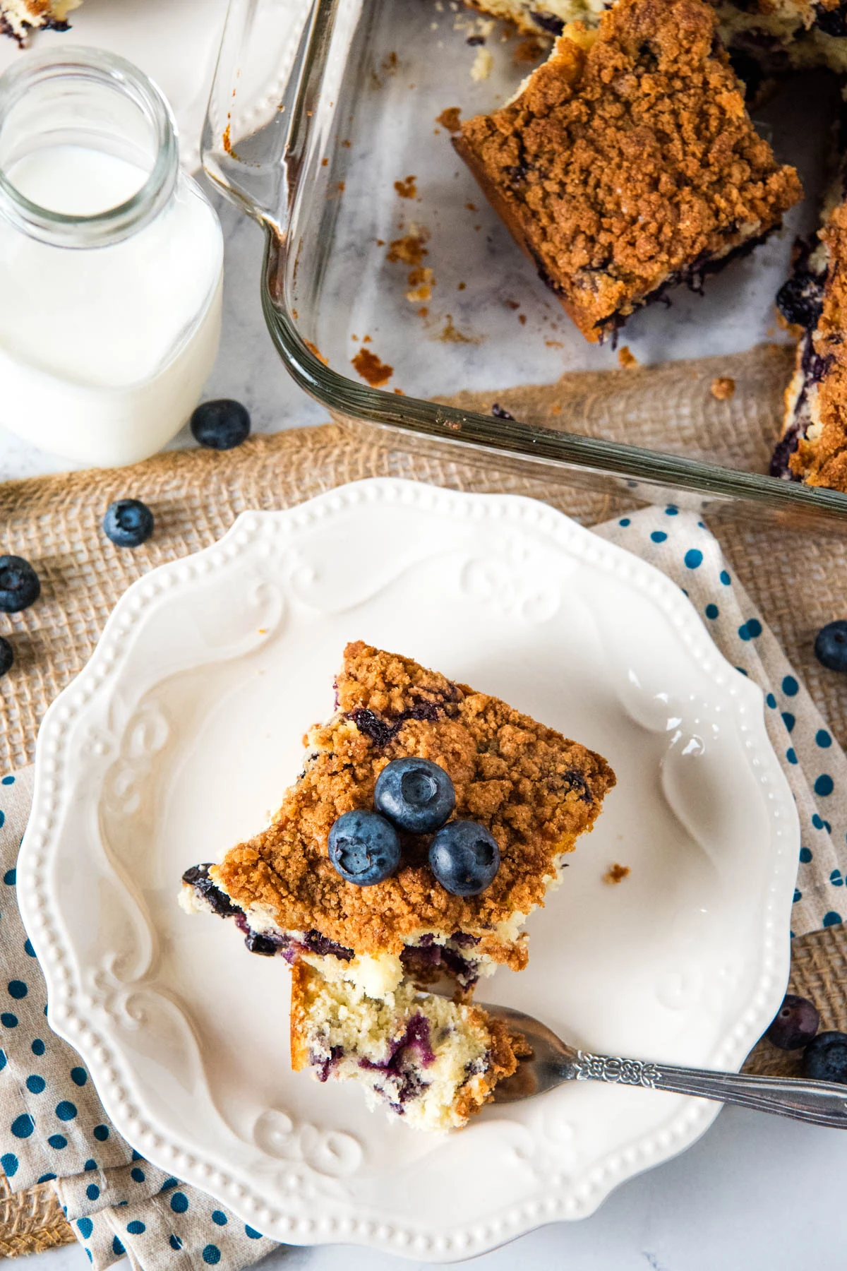 sliced square of blueberry coffee cake with a bite on a small silver fork on a small white saucer on a white and blue polka dot towel next to a large glass baking dish full of cake