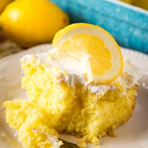 sliced square of lemon Jello poke cake with a forkful of lemon Jello poke cake on a decorative white plate with Cool Whip frosting and a slice of fresh lemon on top