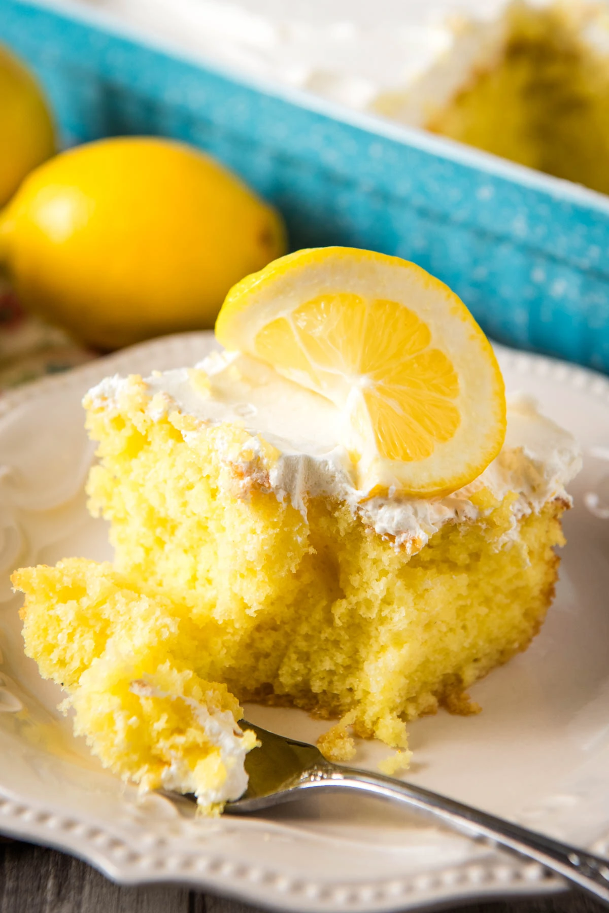 sliced square of lemon Jello poke cake with a forkful of lemon Jello poke cake on a decorative white plate with Cool Whip frosting and a slice of fresh lemon on top