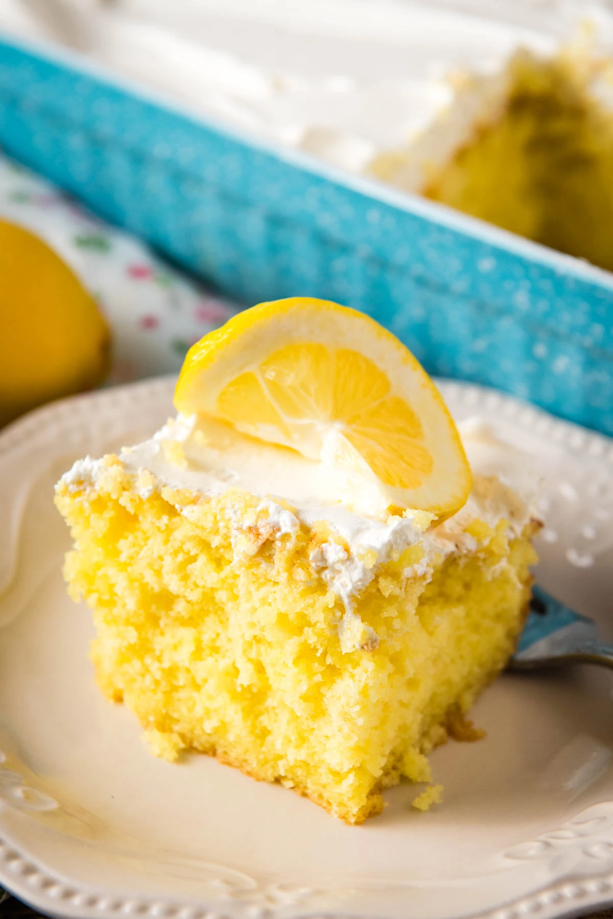 sliced square of lemon poke cake served up on a decorative white plate with Cool Whip frosting and a slice of fresh lemon on top