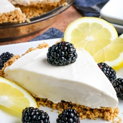 wedged slice of lemon icebox pie with a graham cracker crust on a small white decorative plate with fresh blackberries and lemon slices