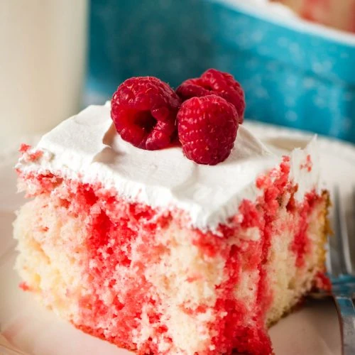 sliced square of raspberry poke cake on a small white saucer with Cool Whip frosting and fresh red raspberries on top