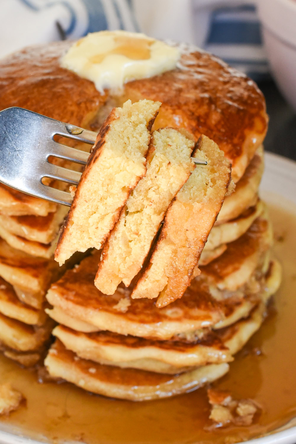 closeup showing a bite of pancakes on a fork with a towering stack of pancakes with maple syrup and butter in the background